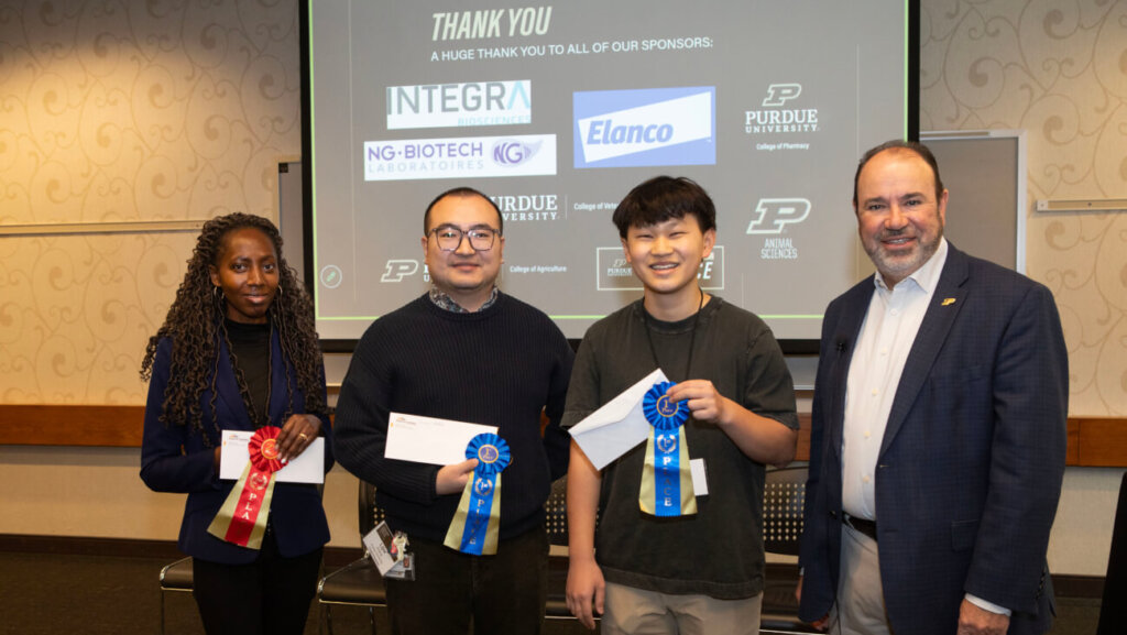 Four people are standing in front of a projector screen. Three of the people are holding ribbons and envelopes.