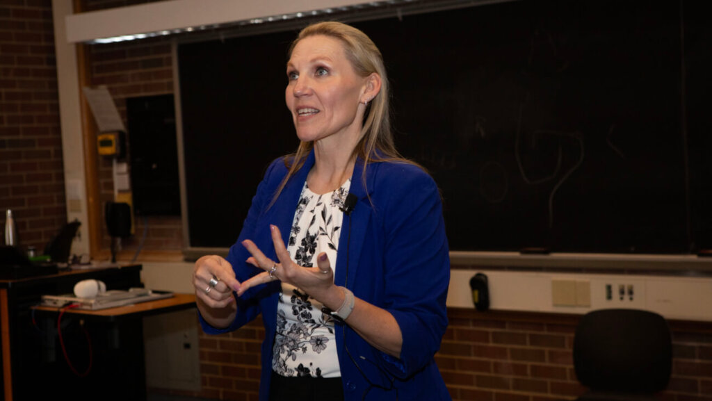 Woman in blue blazer is gesturing with her hands indicating she is mid-sentence. She appears to be doing a presentation. The background is a brick wall.