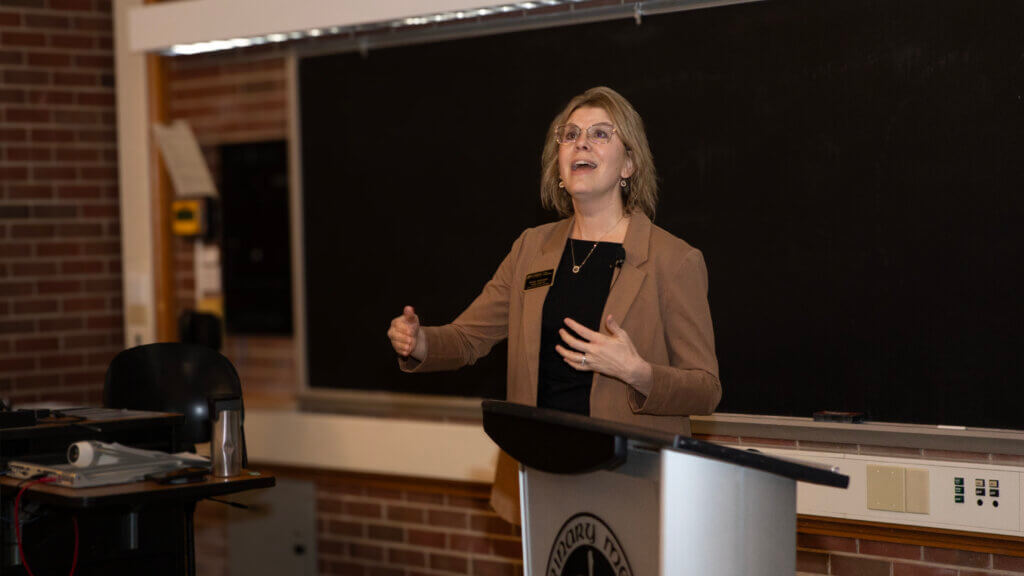A woman wearing a brown blazer is standing at a podium giving a welcoming presentation