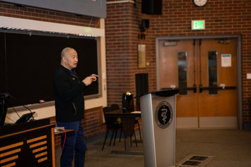 A man wearing black medical scrubs is standing at a podium and gesturing as he is in the middle of a presentation
