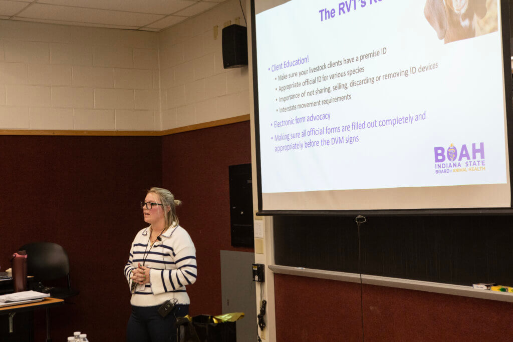 A female is standing in front of a projector screen that discusses content related to the Indiana State Board of Animal Health. The female is wearing a black and white striped sweater.