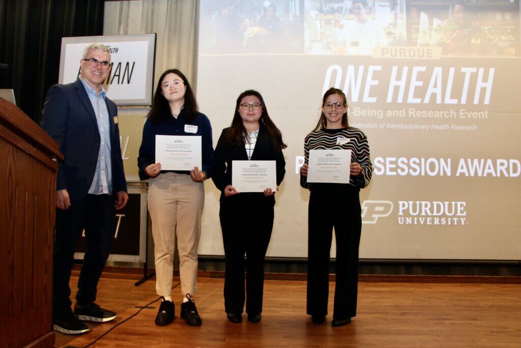 A group of four people are standing on a stage in front of a projector screen that reads "Purdue One Health". Three of the people are holding certificates.