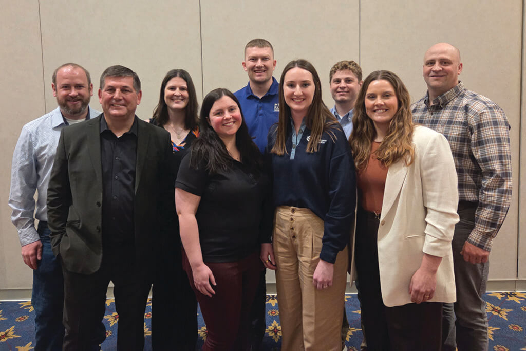 A group of nine people are wearing business attire and are standing in front of a tan wall.