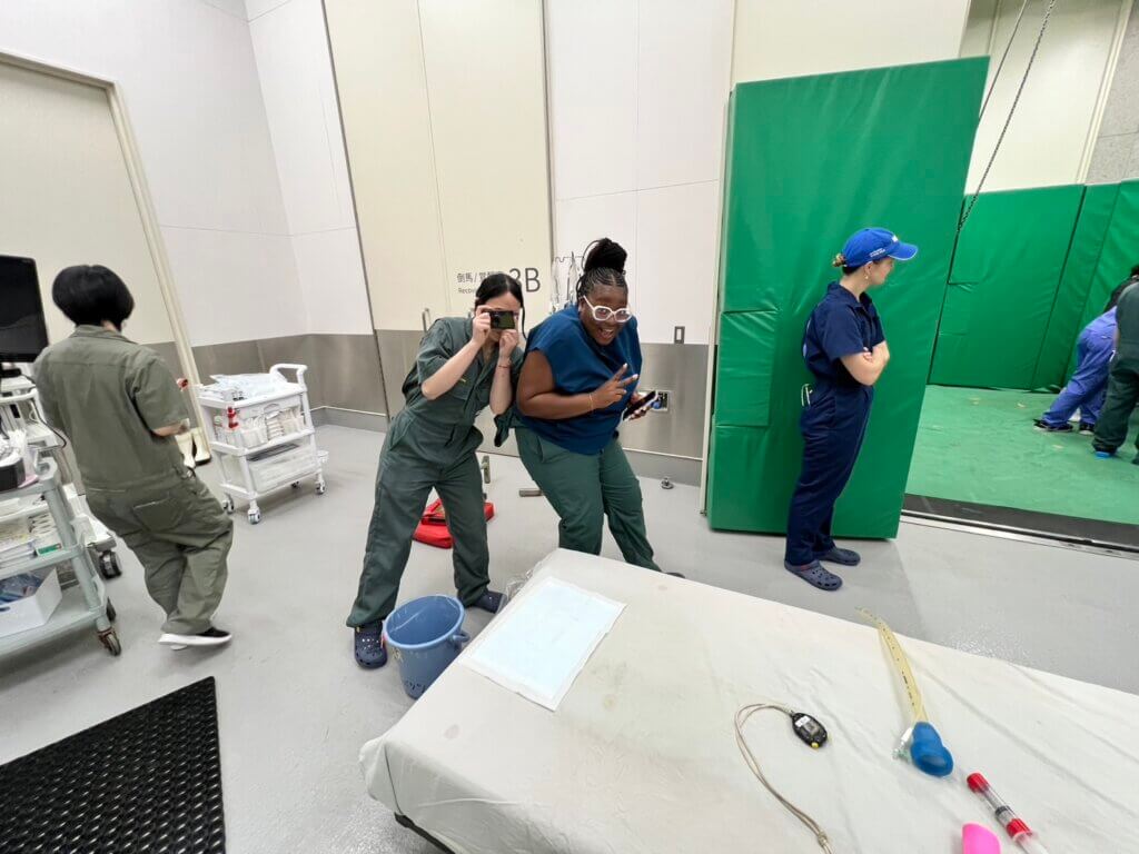 several people are in a padded area of what looks like a medical facility. The main subject of the image are two females in medical attire. The one on the left is holding a camera and is posing toward the viewer as if she is taking a photo.