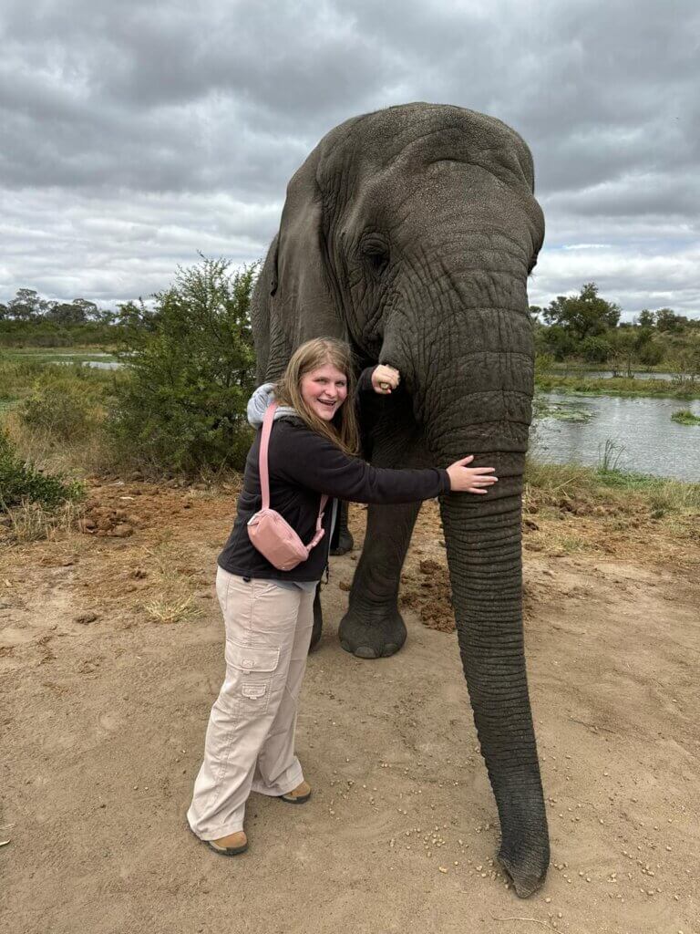 A female is standing with an elephant embracing the animal's trunk. The female is wearing a black shirt and a pink bag.