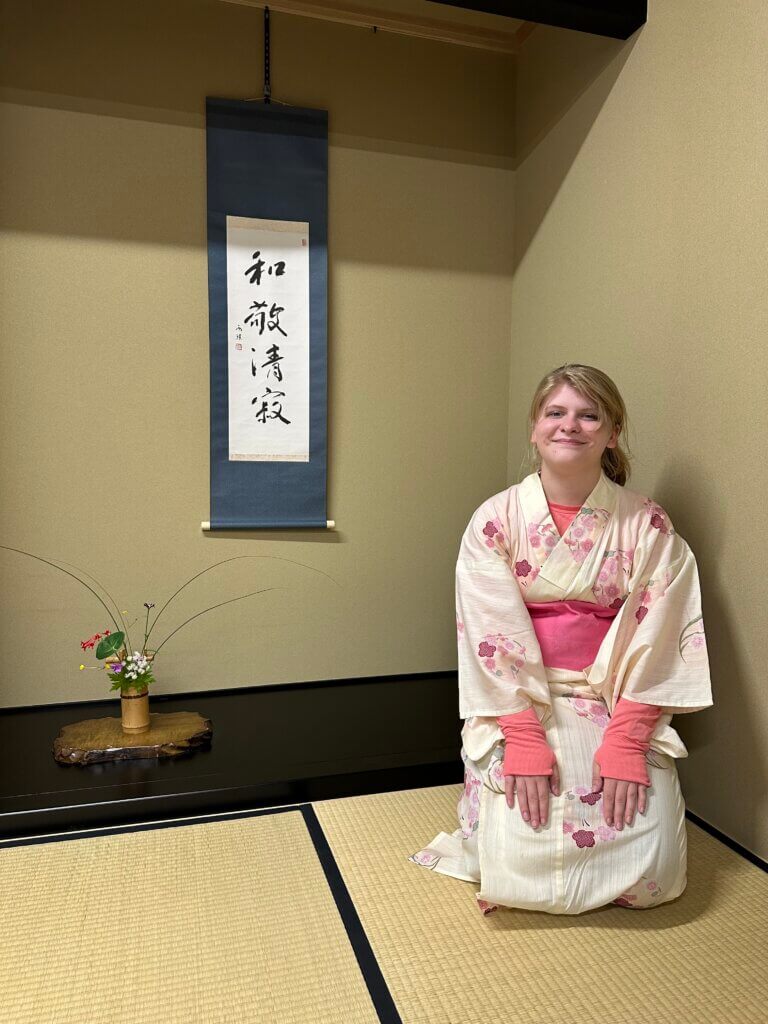 A female is kneeling on the floor of what seems to be a Japanese room. She is wearing a pink oriental piece of clothing