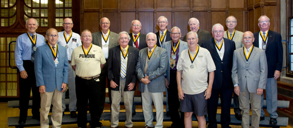 A group of approximately sixteen men are standing in a staggered arrangement for a group photograph. Each of them is wearing a black and gold lanyard.