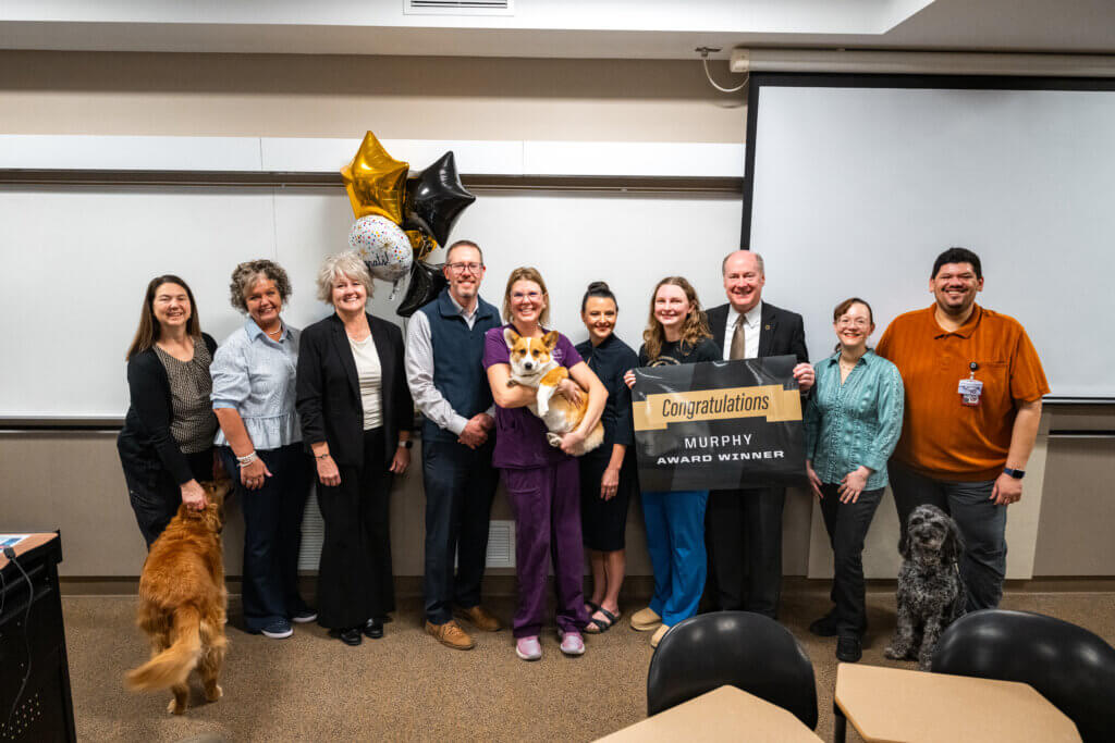 A group of ten people are standing at the front of a classroom in a variety of colored clothing. There are dogs on both sides of the group. There are gold and black balloons, and two people are holding a sign that says "Congratulations Murphy Award Winner"