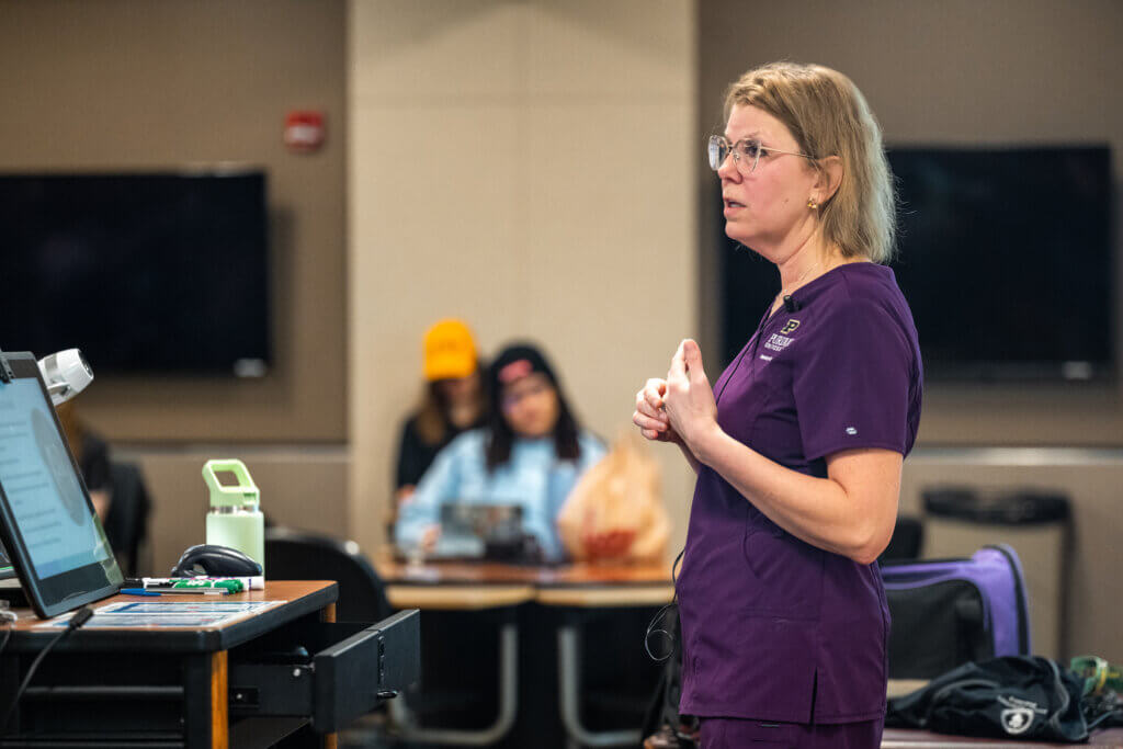 A woman wearing purple medical scrubs is standing and speaking to a classroom of students