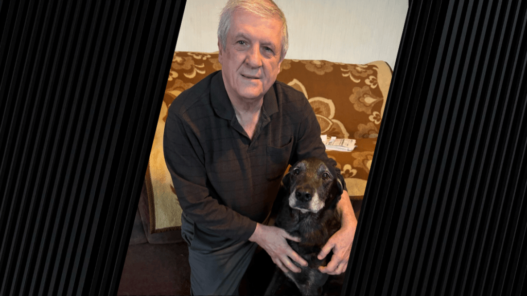 A man wearing a black shirt is kneeling down next to a black dog.