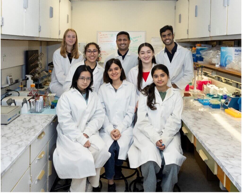 A group of eight individuals are posed for a group photo in a laboratory setting with work benches on both sides of them. Each person is wearing a white lab coat.