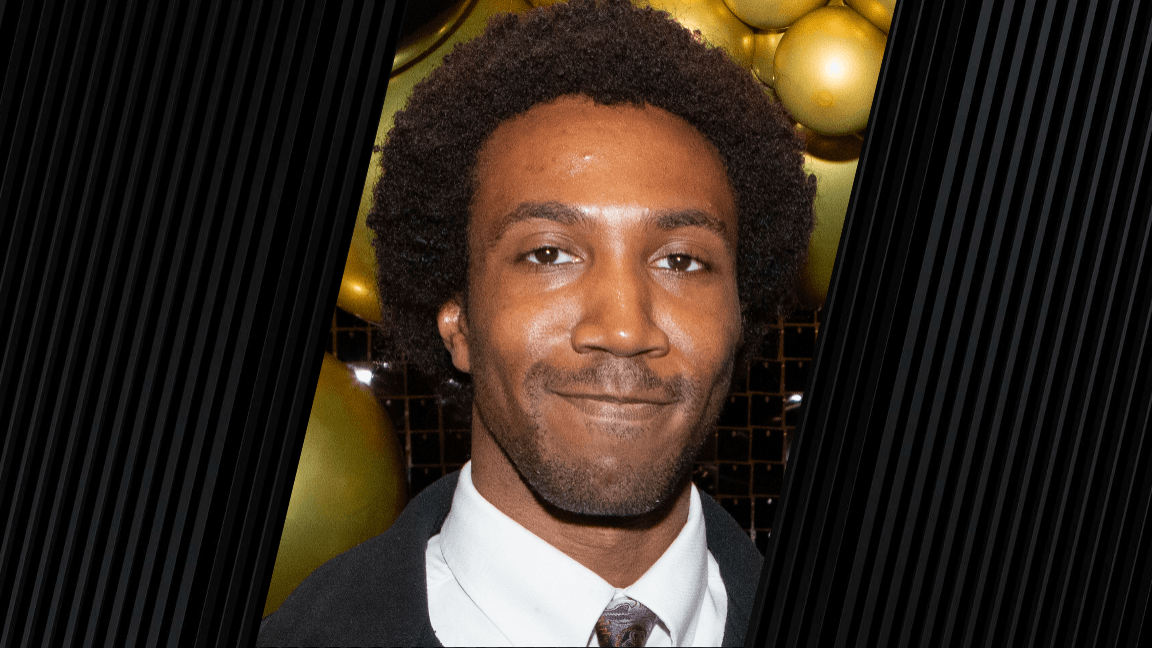 man wearing a black tie, white shirt and black jacket. The background appears to be gold and black balloon.s