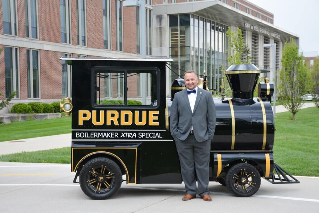 A man stands in front of a "PURDUE" labeled train, and he is wearing a gray suit