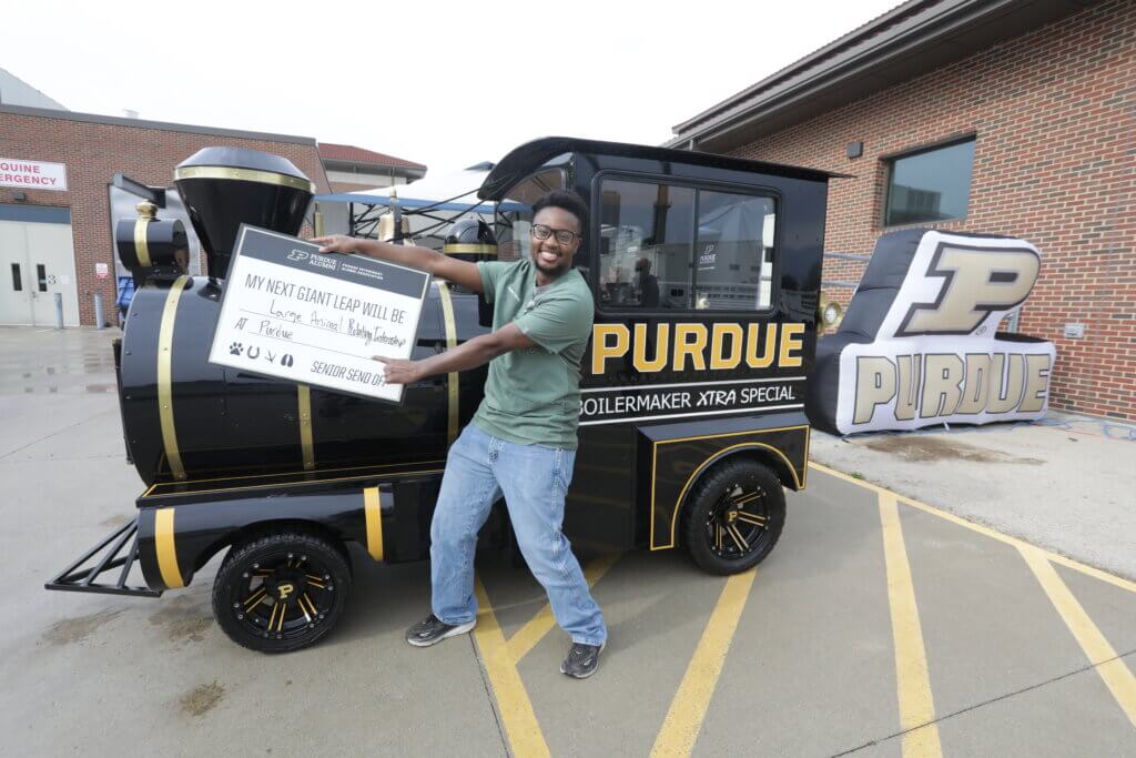A man is standing in front of a locomotive that says "PURDUE". He is wearing jeans and a green shirt