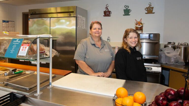 Two people are standing behind a food service counter. One of them is wearing a short-sleeve gray shirt and the other is wearing a black sweater.
