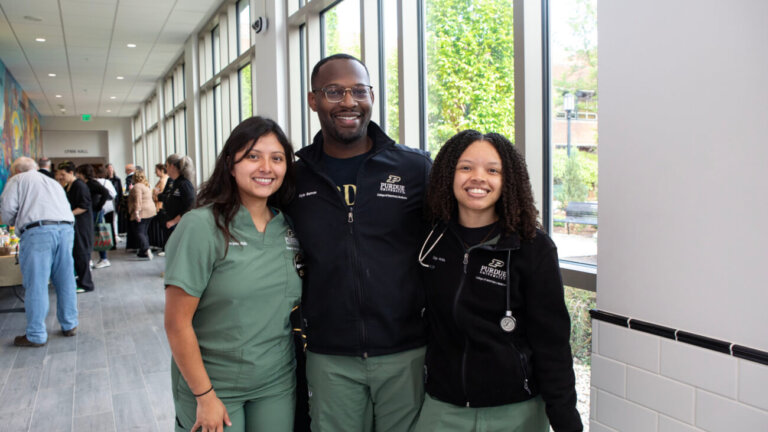 Three individuals are standing in front of a window. All three are wearing olive green medical scrubs.