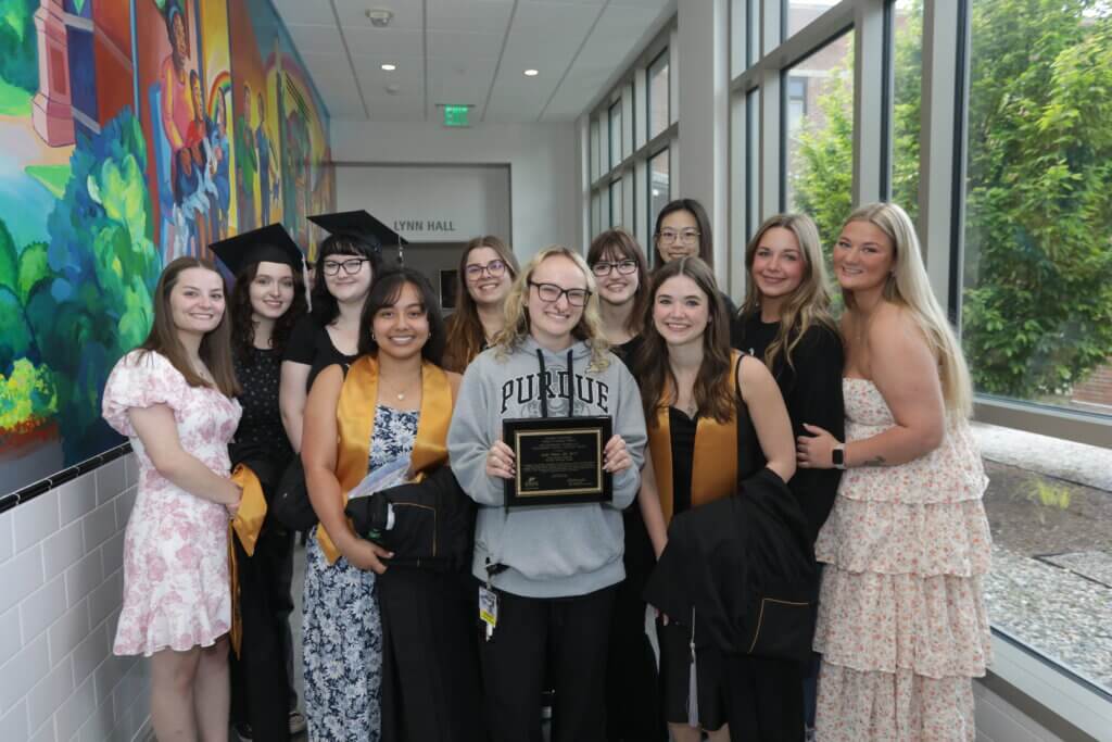 A group of people are standing in the middle of a long corridor with a colorful mural painted on the wall. The person in the middle of the group is holding an award.