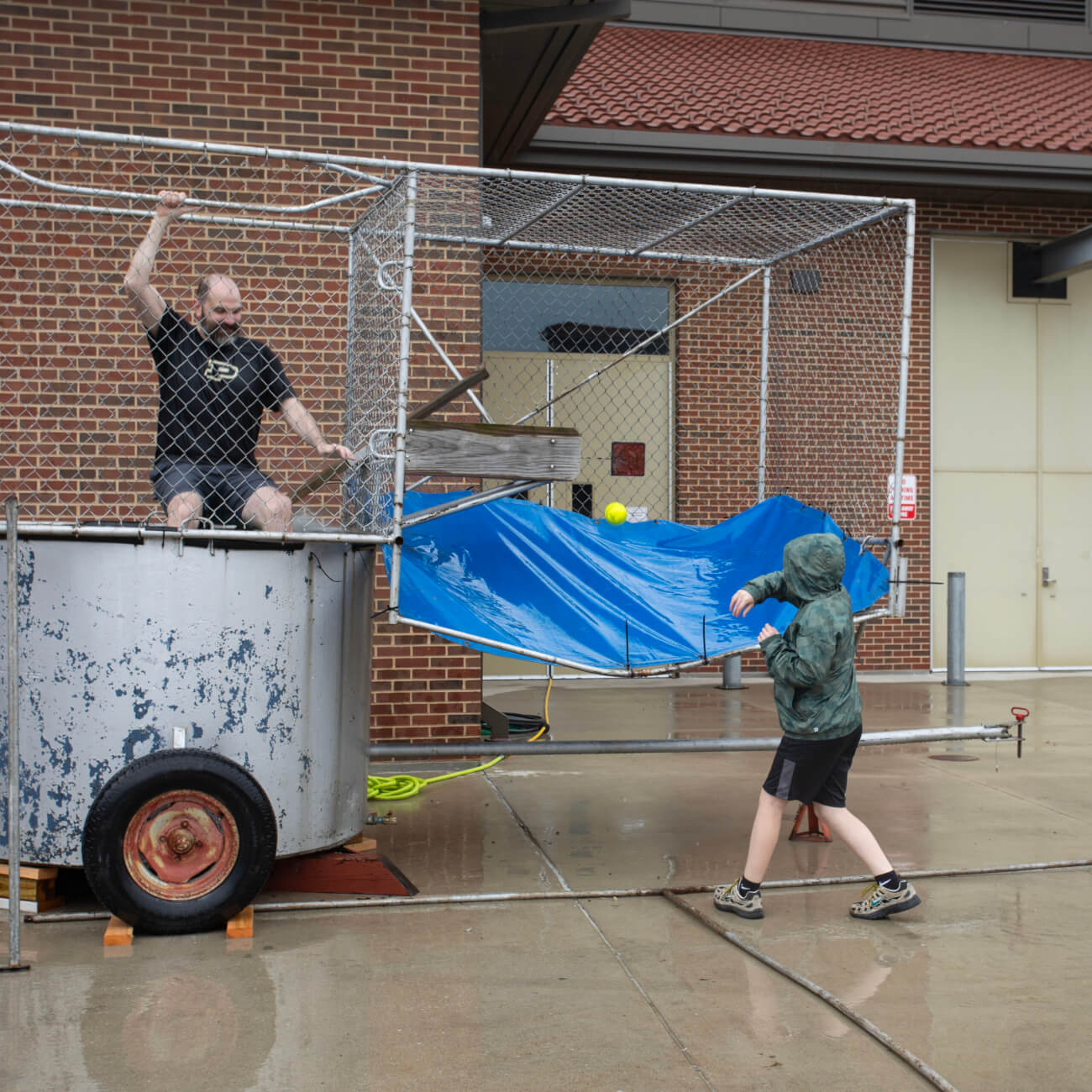a dunk tank is stationed outside a brick building. A man is sitting inside the dunk tank while a child is outside making an attempt to initiate the dunking