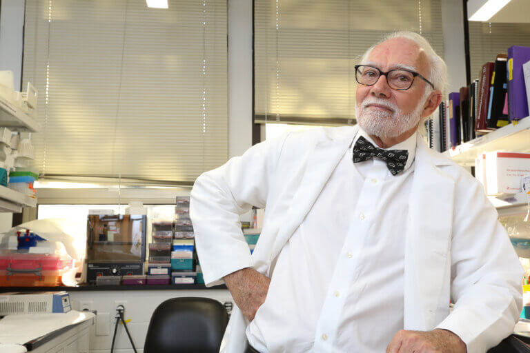 A man is wearing a white shirt, white jacket, and black bow tie. The man is standing in a laboratory setting and leaning on a counter.