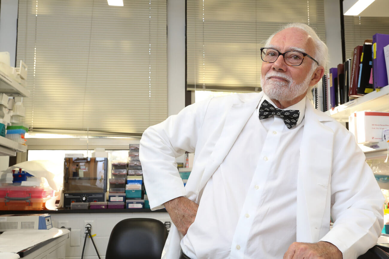 A man is wearing a white shirt, white jacket, and black bow tie. The man is standing in a laboratory setting and leaning on a counter.