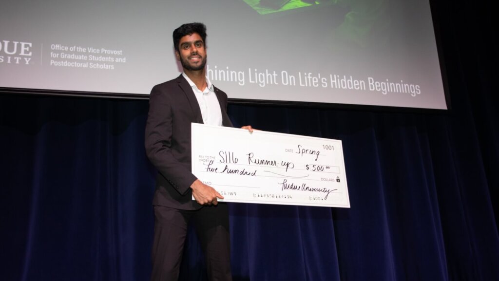 A man is standing on a stage holding a large check that reads "SII6 Runner-Up, $500". The man is wearing a dark gray suit and is standing in front of a blue stage curtain.