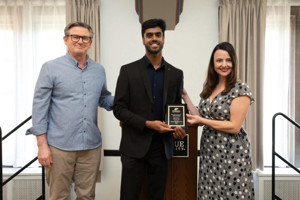 Three individuals are standing in honor of an award. Two men are on the left and center, and a woman is on the right. The man in the center is holding the award.
