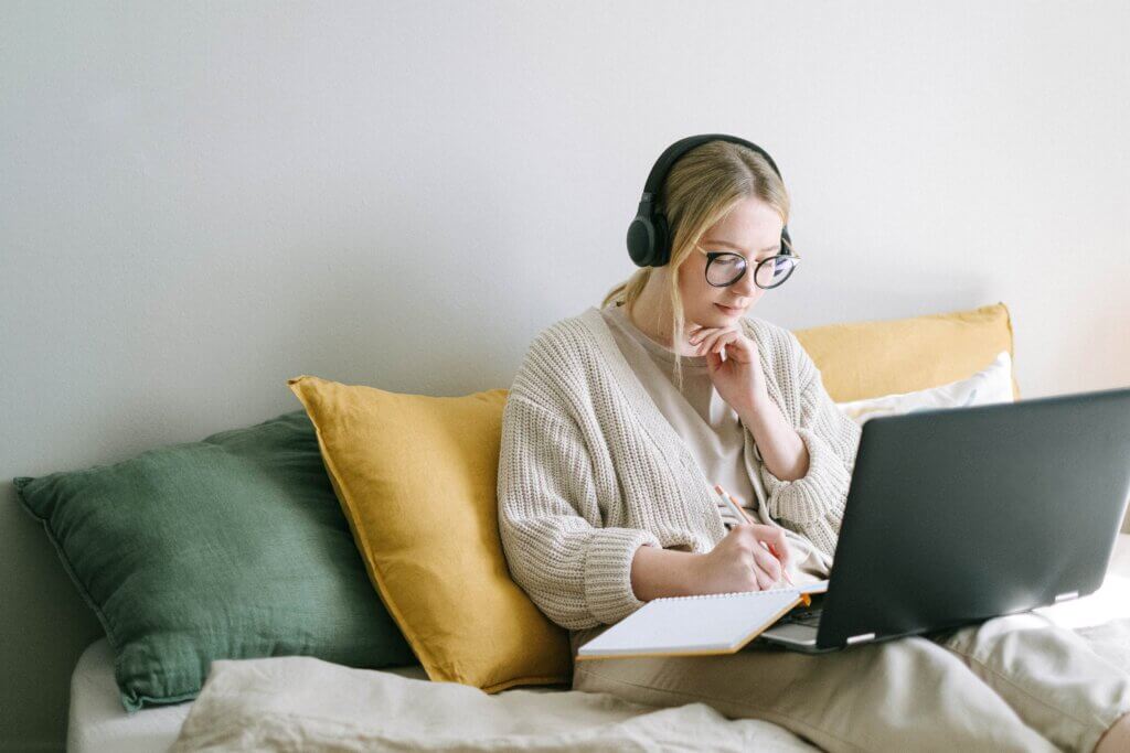 A young woman wearing glasses and over-ear headphones sits on a bed, focused on her laptop while writing in a notebook.