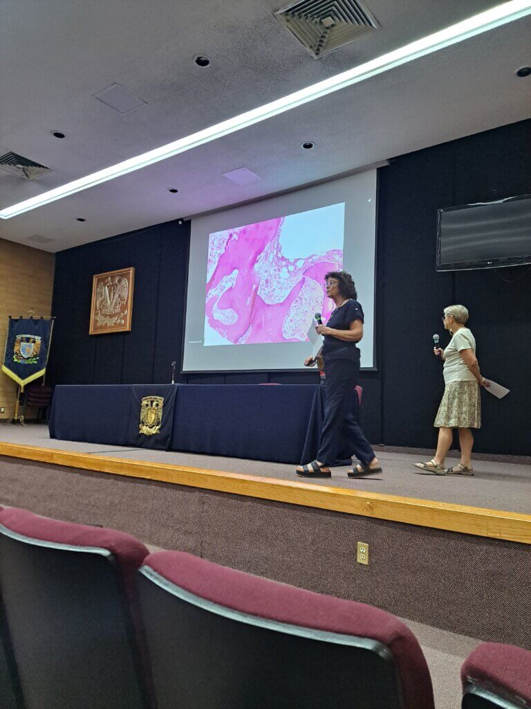 Two women are giving a lecture from a stage. A microscopic image is displayed on a projector screen as they are speaking to an audience.