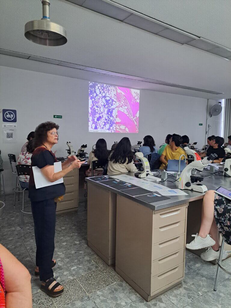 A woman is teaching a group of students in an indoor setting. She is standing while the students are sitting, and she is using a projector screen to display a microscopic image.