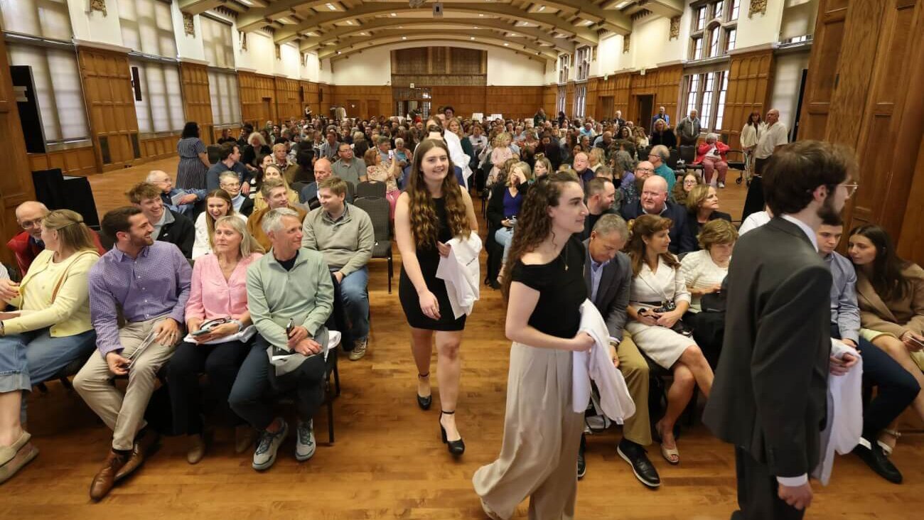 A large audience is seated in a ballroom. There is a center aisle with students walking down it.