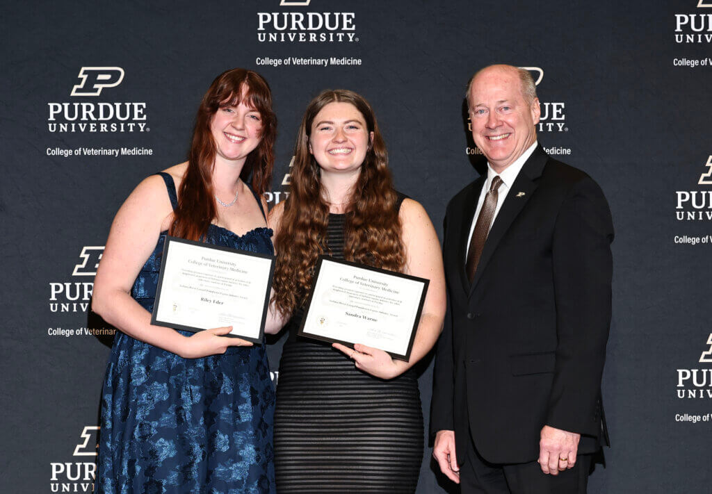 Three people are standing in front of a Purdue University-themed backdrop. The two women on the left are wearing dresses and holding awards and the man on the right side of the image is wearing a black suit.