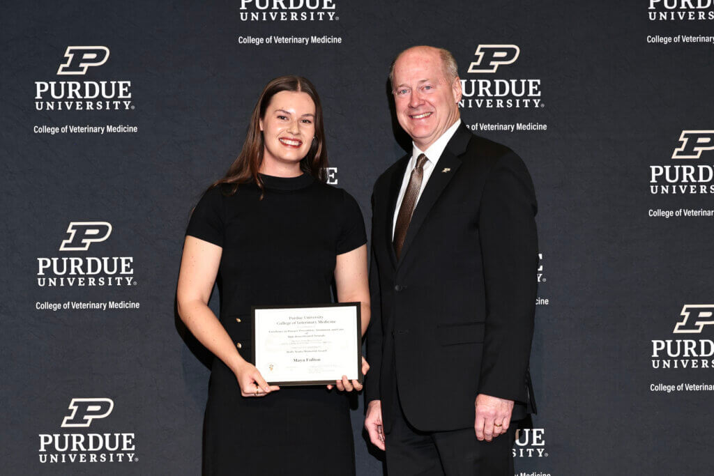 A female is on the left side of the image and is wearing a black dress holding an award. A man is standing next to her on the right and is wearing a black suit.
