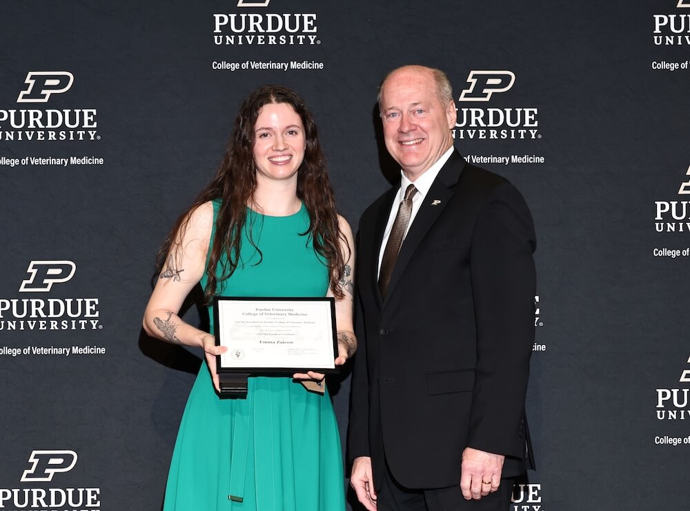 A woman is standing on the left side of the image and is wearing a green dress and holding an award. A man in a black suit is standing next to her on the right side of the image