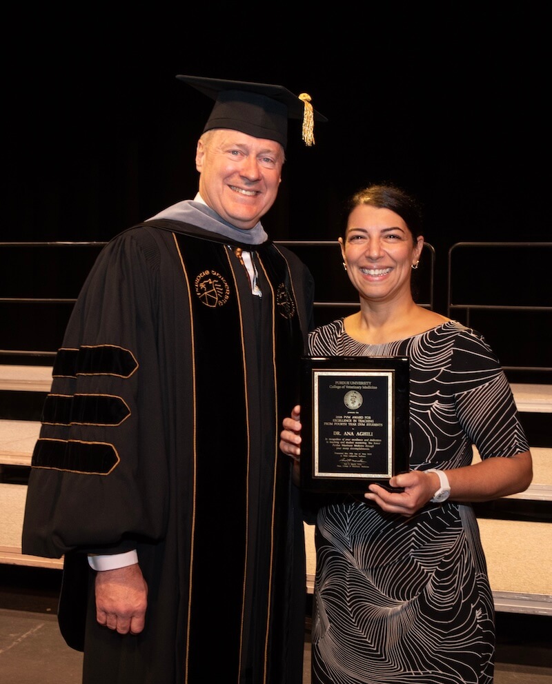Two people stand next to each other in graduation and business formal attire. The woman is standing on the right and is holding an award, and the man on the left is wearing graduation ceremony attire.