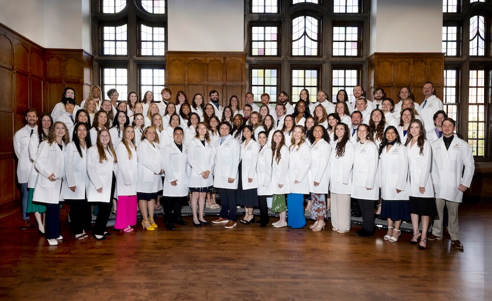 A large group of young adults are positioned on risers and all wearing white medical coats.