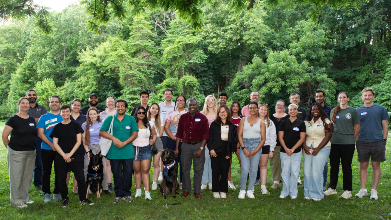 A group photo featuring both humans and dogs and set in a natural landscape with vibrant green trees in the background