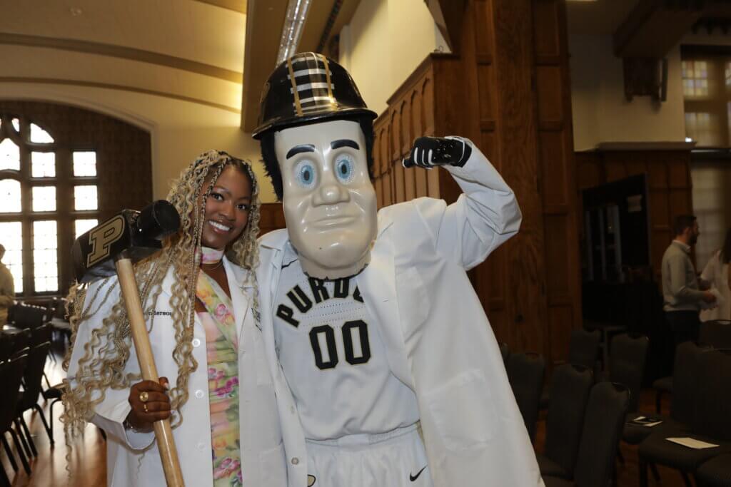 The Purdue University mascot 'Purdue Pete' is posed flexing his bicep next to a graduated veterinary student who is wearing a white coat and holding a Purdue University-branded hammer