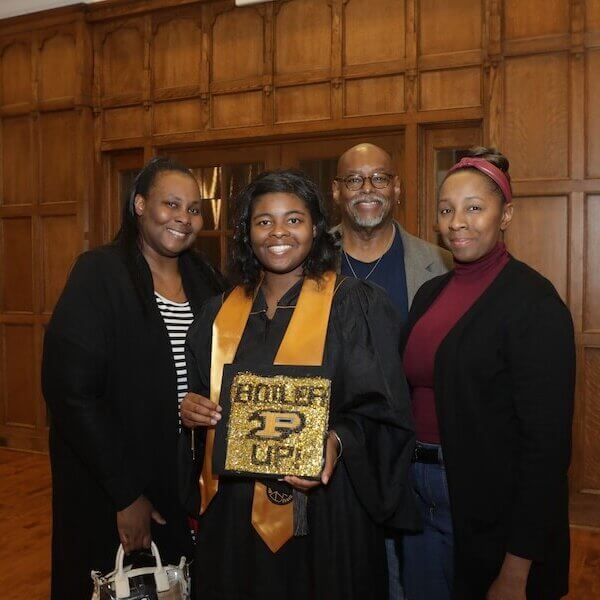 A group of four people are gathered for a group photo of a graduate. The graduate is holding a sign that reads "BOILER UP!"
