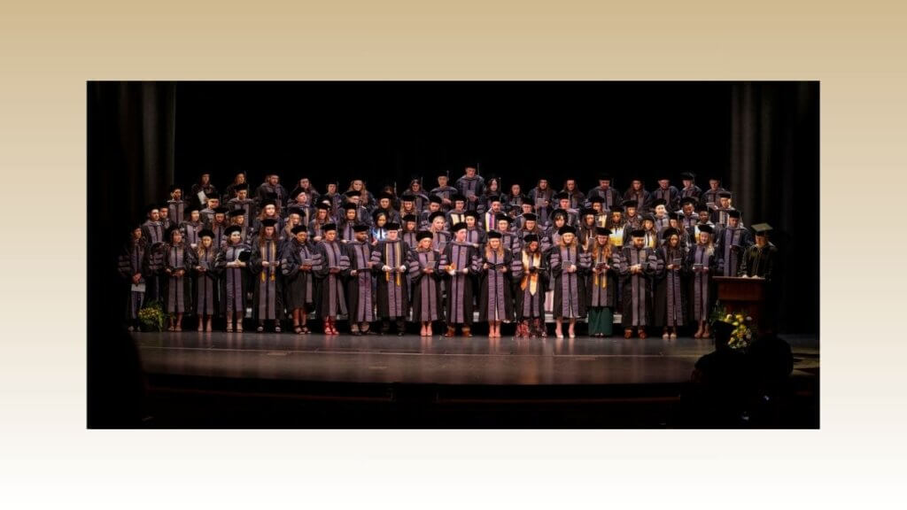 A large group of graduating students are standing on stage wearing graduation caps, gowns, and sashes