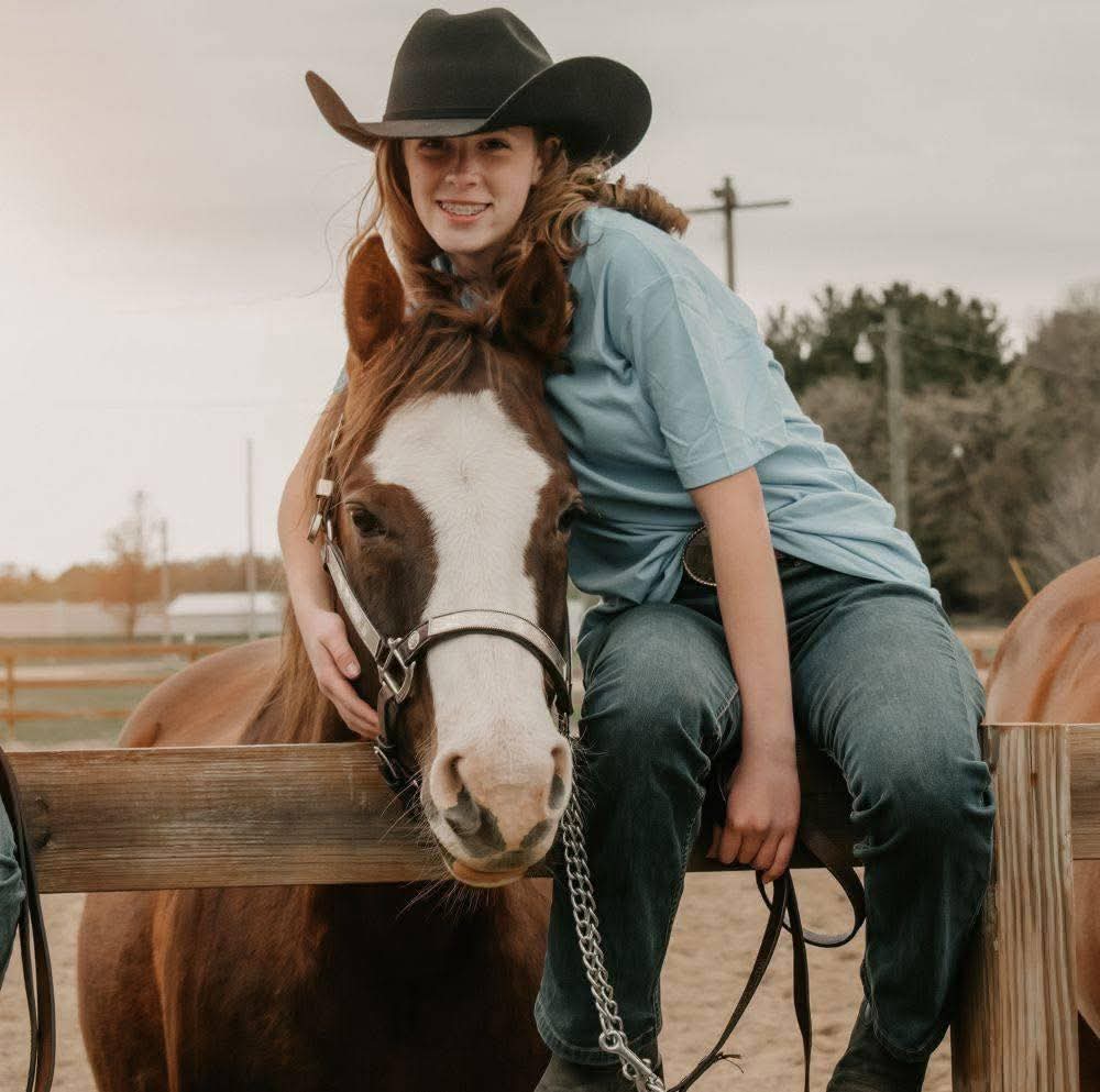 A female is sitting on a fence next to a horse. The human is wearing a blue shirt and black cowboy hat. The horse has brown-red coloring and white fur on her nose and face.