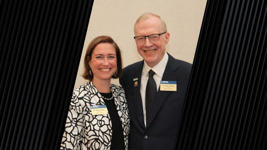 A woman and man stand next to each other smiling. Both are wearing suit jackets. The woman is on the left side of the photo and the man is on the right side of the photo.