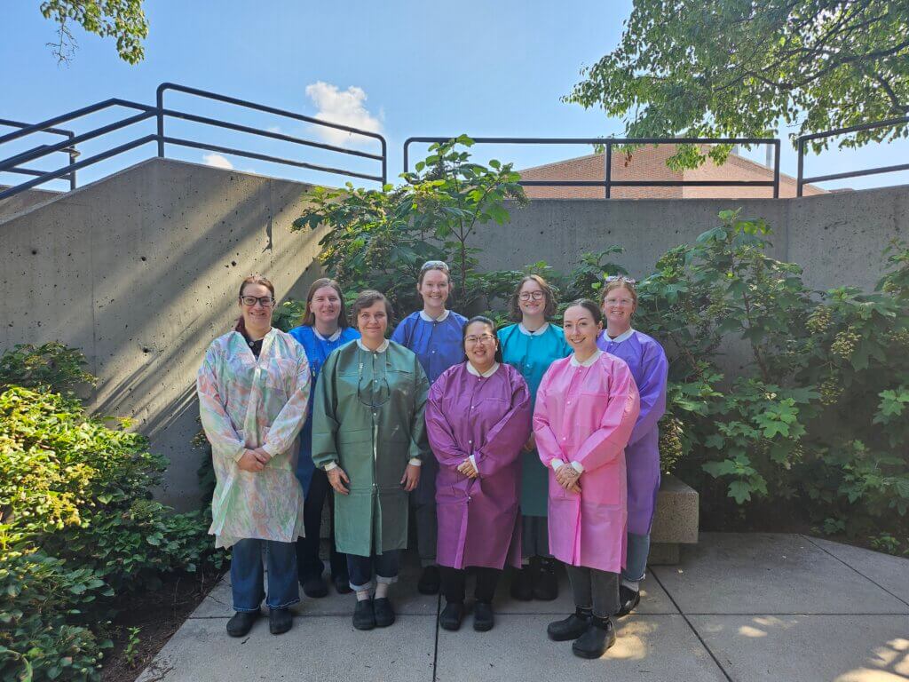 A group of women are standing outdoors in front of a concrete wall and small trees. The women are wearing laboratory coats of various colors.