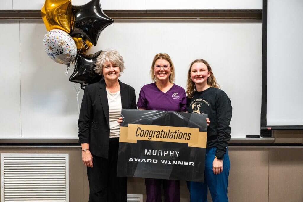 Three women are standing next to each other as an award is presented. The woman in the middle is holding a sign that says "Congratulations MURPHY AWARD WINNER"