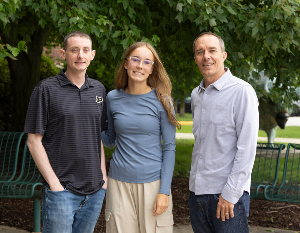 Three figures are standing in front of an outdoor, natural backdrop. The male on the left is wearing a gray shirt with Purdue branding, the female to his right in the photograph is wearing a blue shirt, and the man on the far right of the photo is wearing a gray button-up shirt.