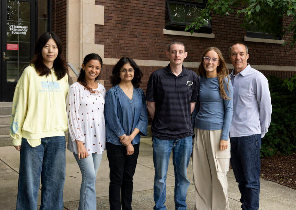 Six people of various ages and ethnicities stand in front of a brick building. They are all wearing business casual attire and smiling at the camera.
