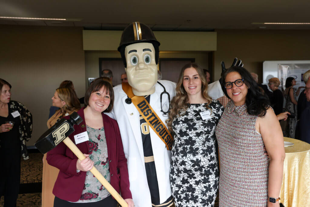 Four figures standing next to each other, with the second from the left wearing a mascot costume of Purdue Pete. The people are dressed in business formal attire