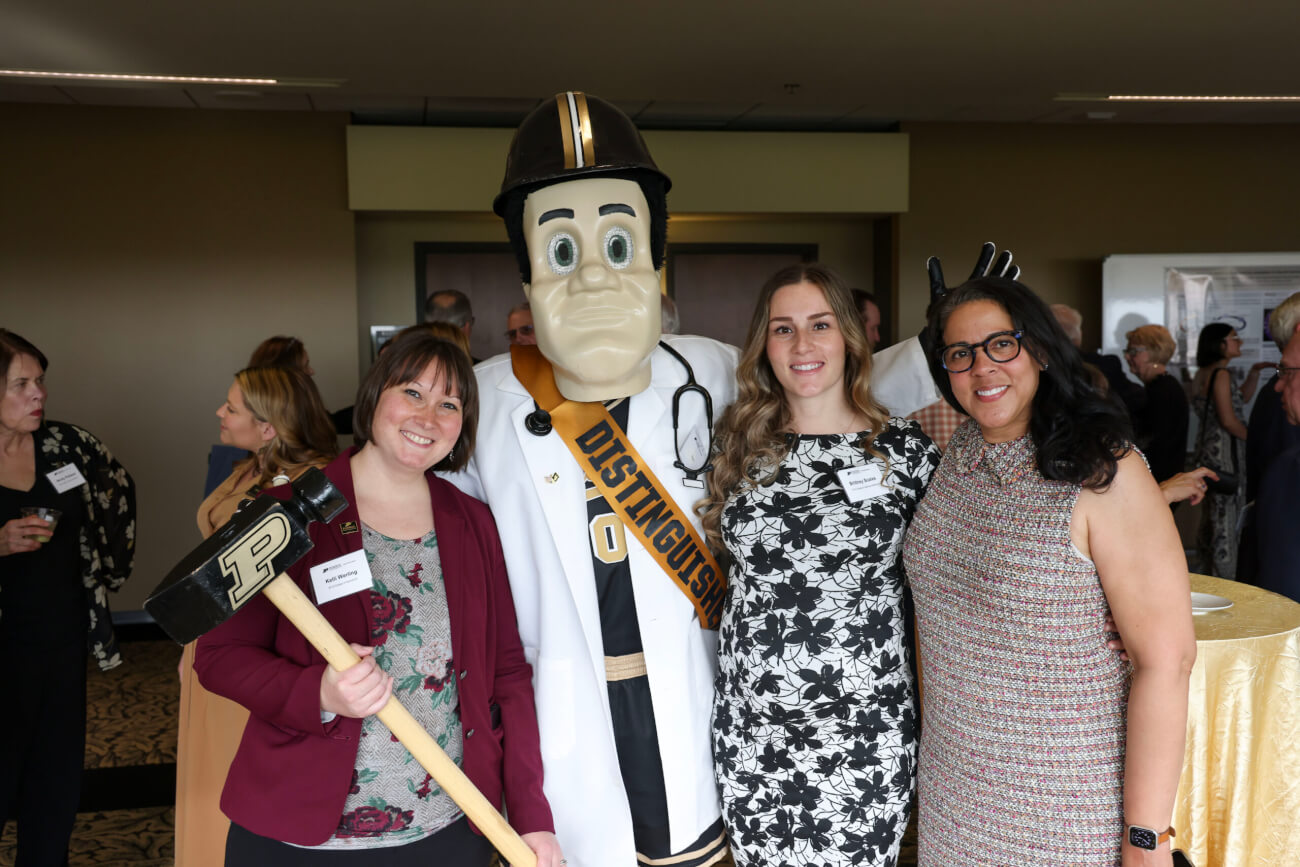 Four figures standing next to each other, with the second from the left wearing a mascot costume of Purdue Pete. The people are dressed in business formal attire