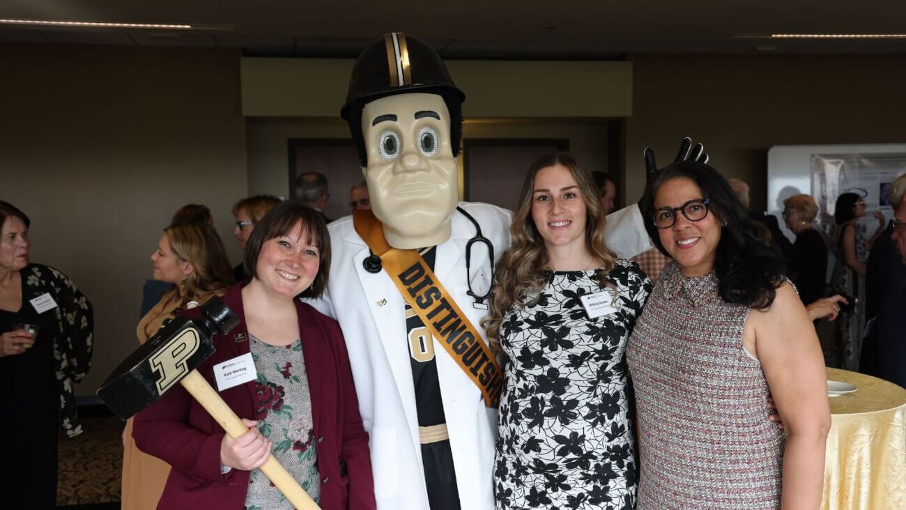 Four figures standing next to each other, with the second from the left wearing a mascot costume of Purdue Pete. The people are dressed in business formal attire
