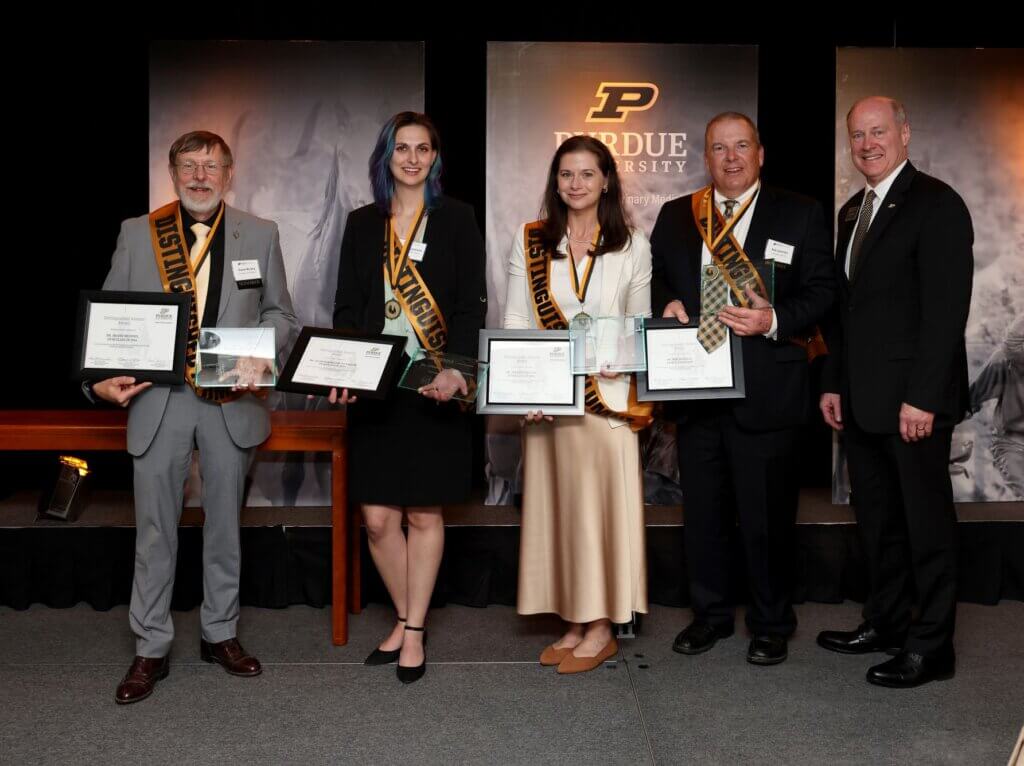 Five people are standing next to each other on a stage, and the left-most four people are holding awards and wearing sashes.