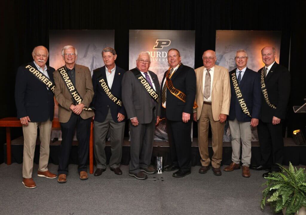 A group of eight men are standing on stage, with sashes that say "former honoree"